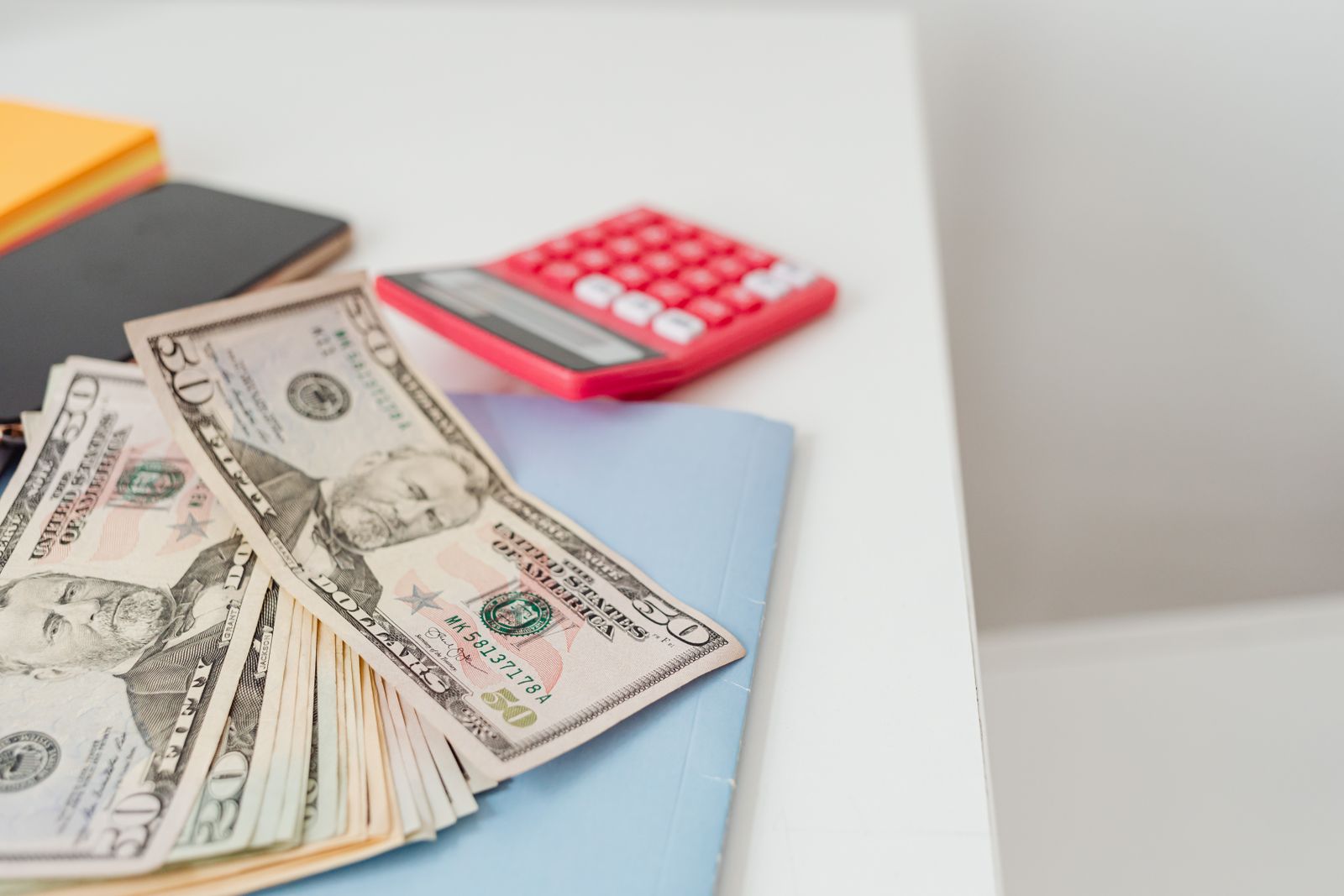A fan of banknotes resting on a notebook beside a red calculator on a clean white desk.