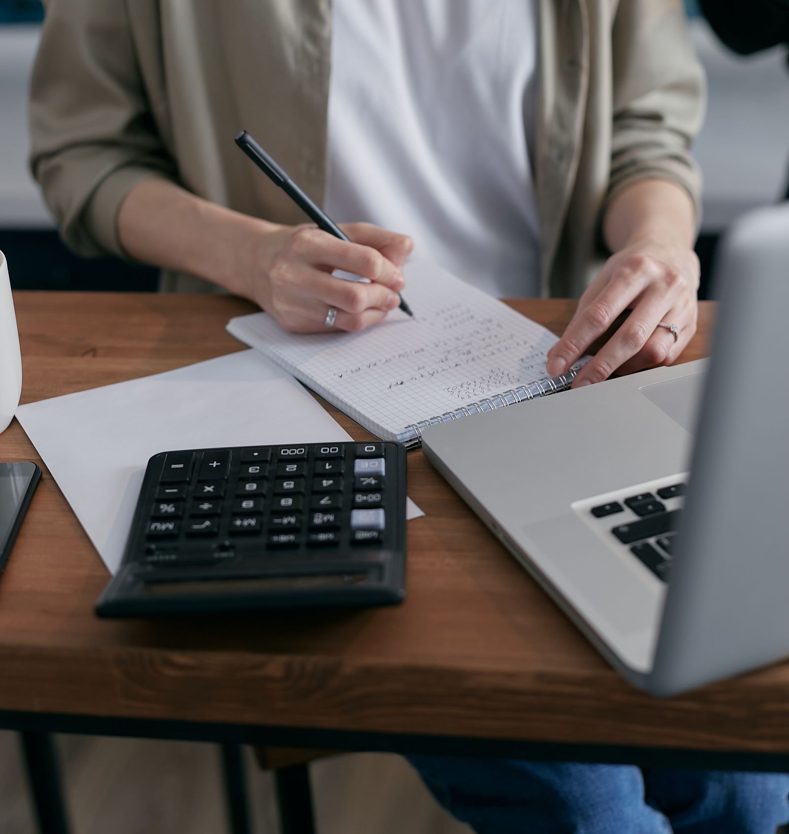 A person's hands writing figures in a notebook beside a calculator and laptop, planning their finances at a desk.